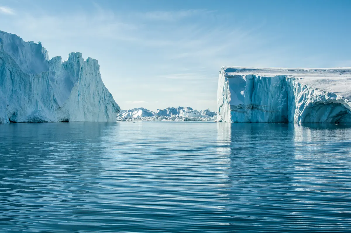 Des icebergs se reflètent dans une mer calme, sous un ciel bleu.